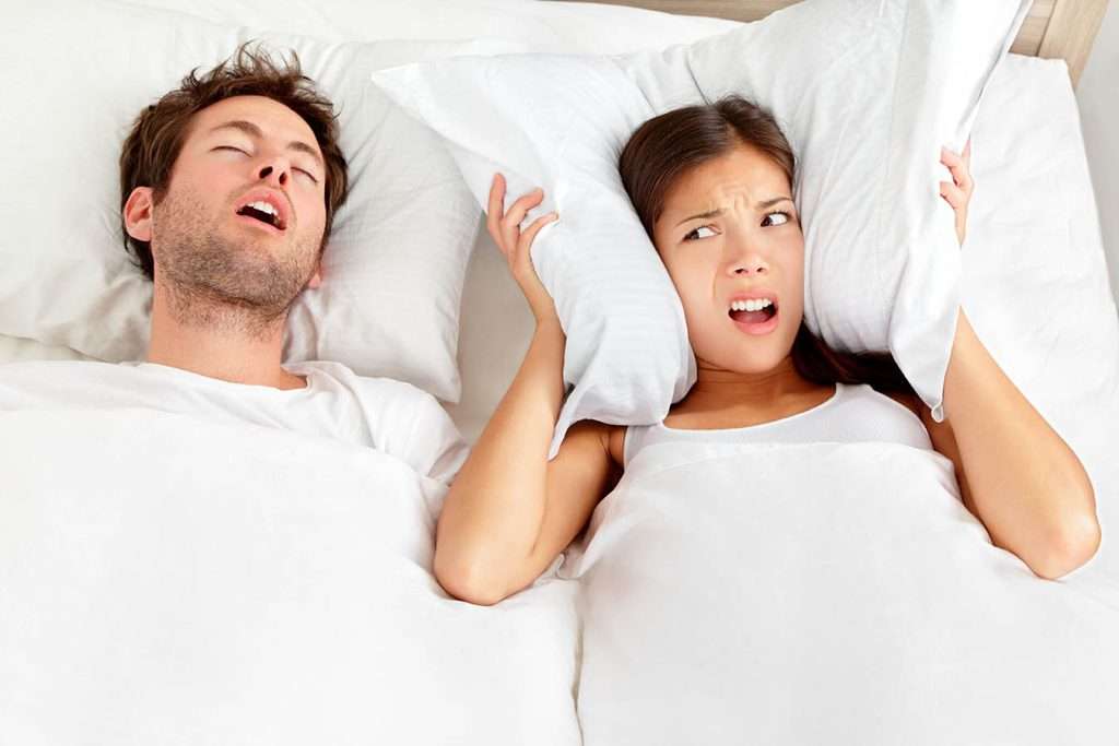 Snoring man with Sleep Apnea. Los Angeles couple in bed, man snoring and woman can not sleep, covering ears with pillow for snore noise, wondering what is causing his sleep apnea.